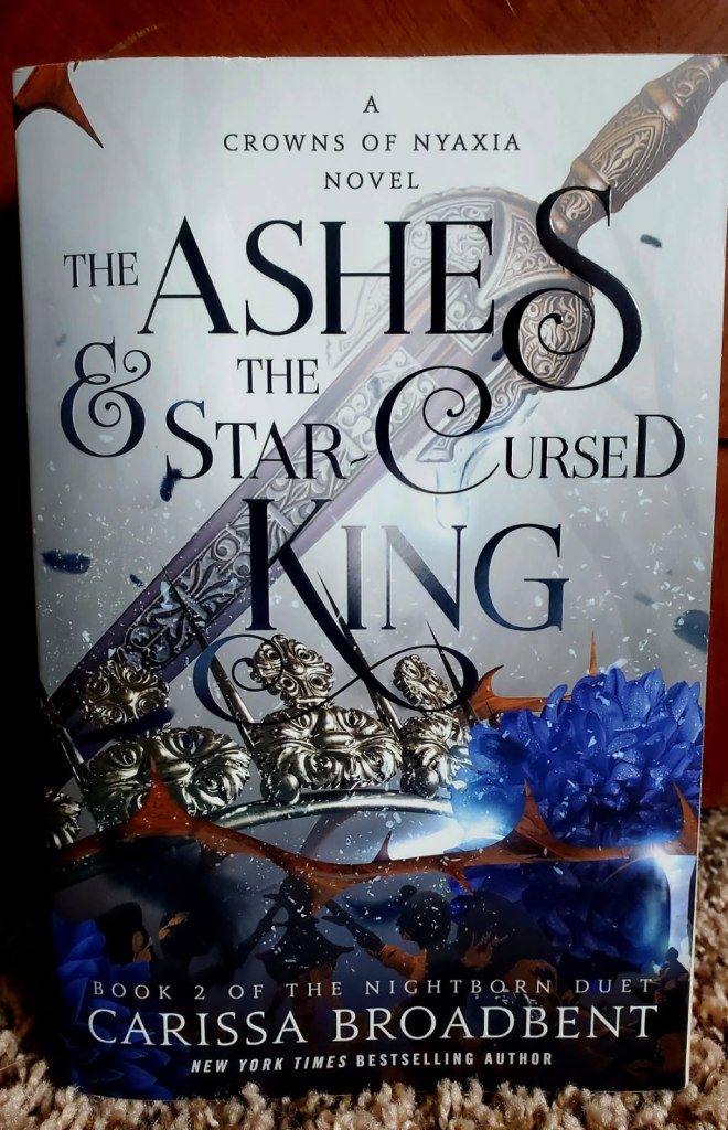 Image is a book cover for The Ashes & The Star-Cursed King by Carissa Broadbent. The book shows a sword with a crown and blue flower next to it. 