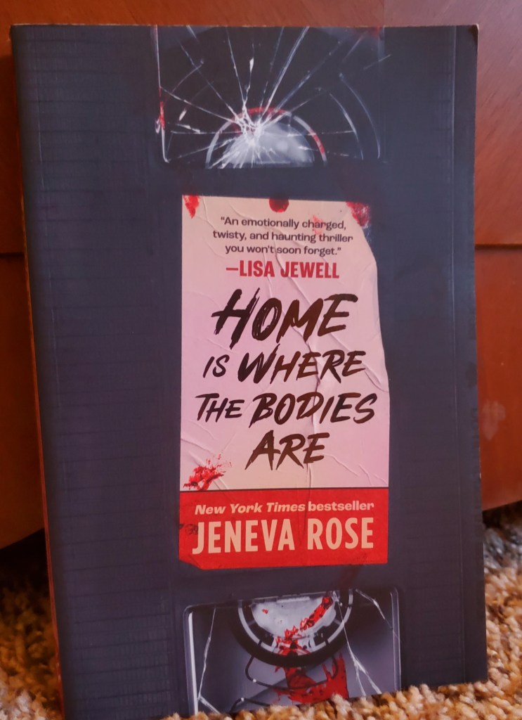 Image is a book cover for Home Is Where the Bodies Are by Jeneva Rose. It's a VHS tape that's cracked and the title of the book looks like a post it note with blood on it. 