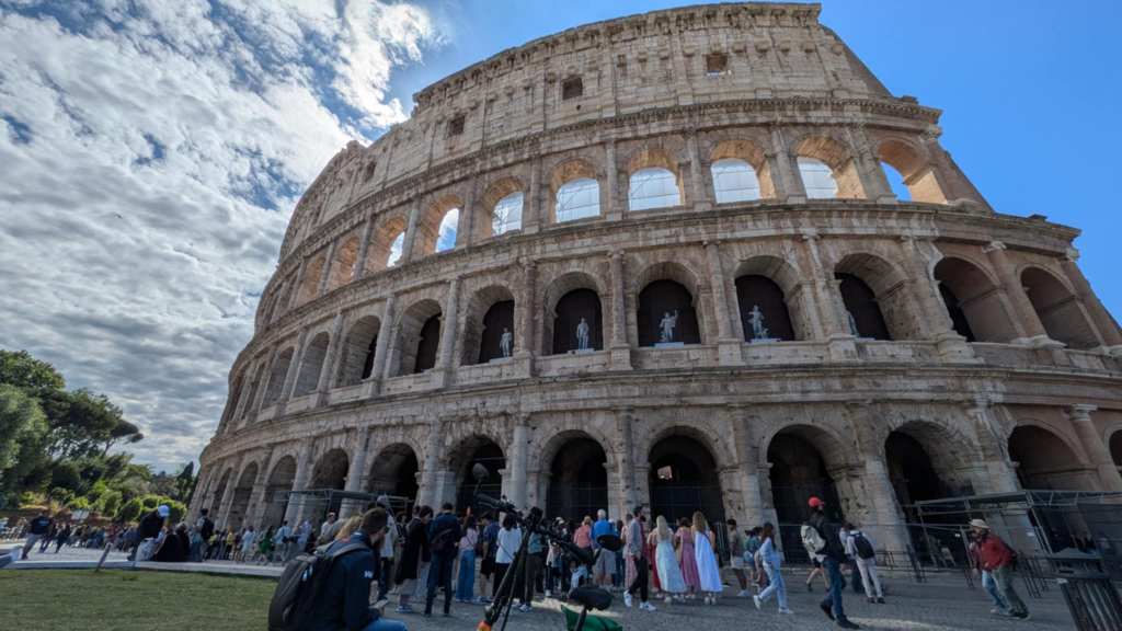Image is a close up view of the Colosseum. The sky is blue with white clouds on the left of the photo. It shows three layers to the colosseum with the third layer collapsing. In the second row, there are figures that can be seen through the slots of the colosseum. There are people outside the colosseum looking at it. 