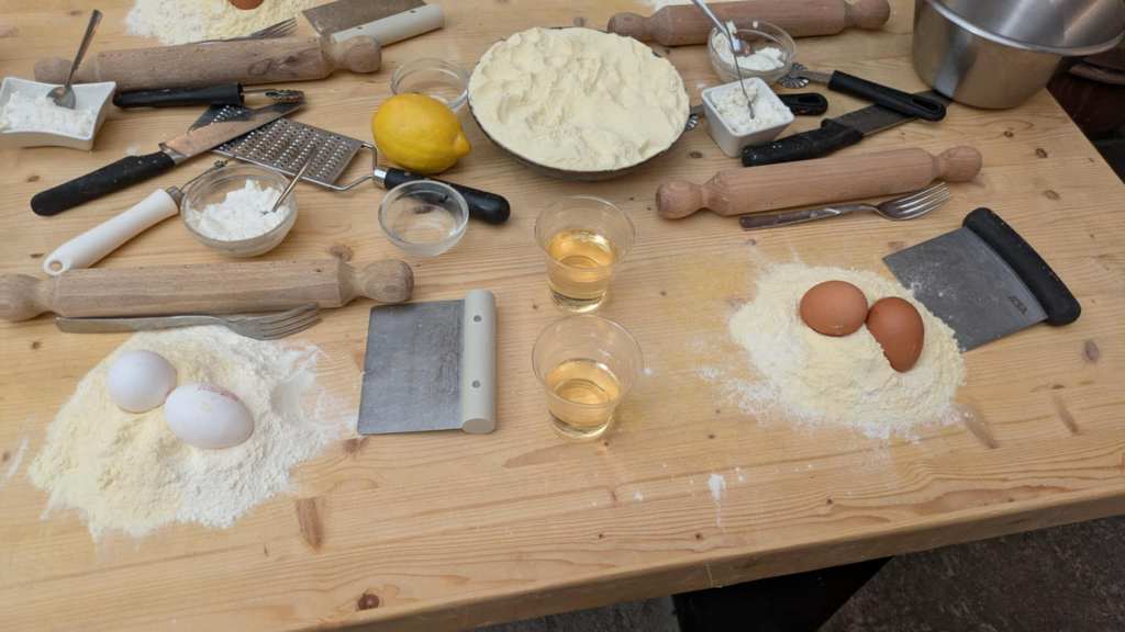 Image shows a wooden table with flour and two eggs on top. There are several tools and dishes with cheese in them. Lots of rolling pins are nearby. 