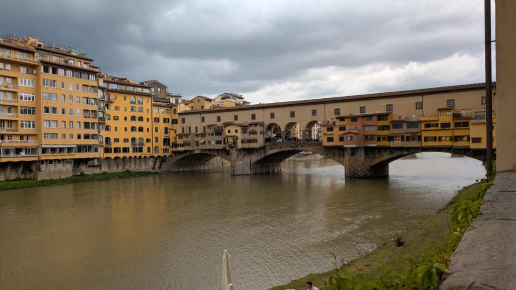 Image is a photo of a medieval stone arch bridge. There are several buildings that jet out of the bridge in yellow and orange. Those are the shops on the bridge. 
