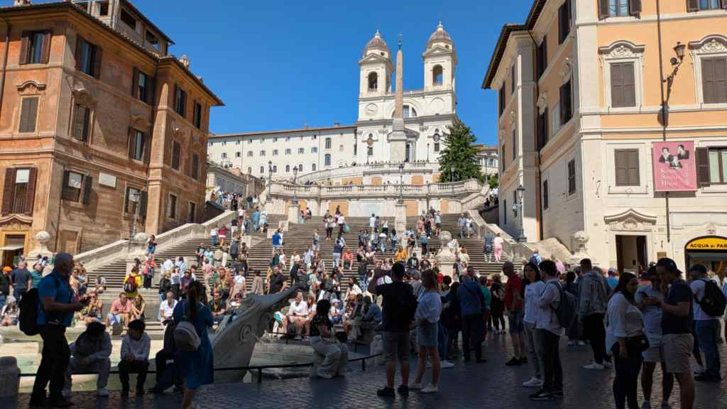 Photo is of the Spanish Steps. There are steps farther away in this photo with a fountain off to the left. There's a white cathedral type building with two pillars / domes at the top. To the left and right of the spanish steps are two Italian buildings with beige, white, and brown stone to them. 