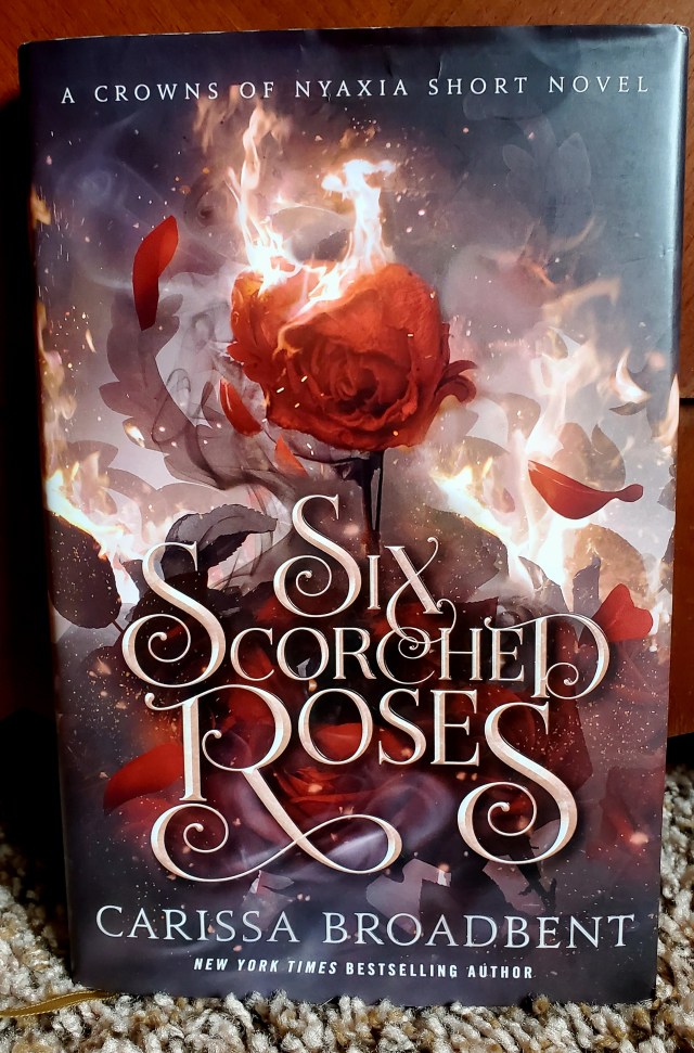 This is a book cover for the book - Six Scorched Roses by Carissa Broadbent. The cover shows a rose on fire. In the background, there's a big fire with rose petals falling on the ground. 