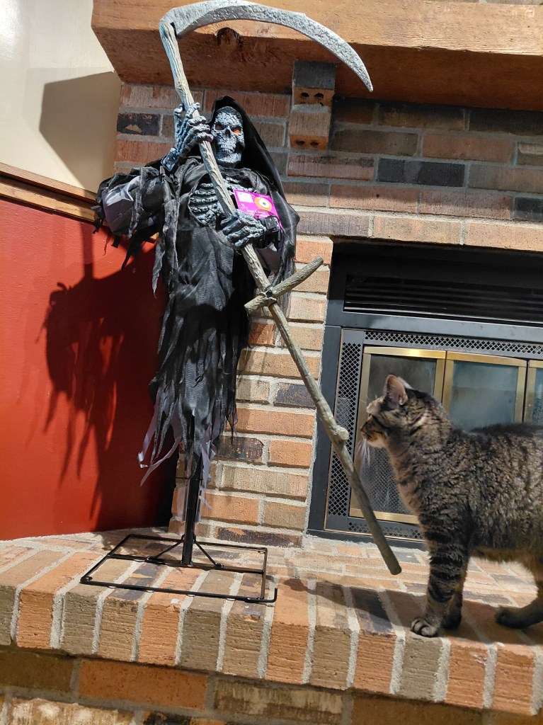 Image is a photo of a Grim Reaper holding a scythe. It's an outdoor Halloween decoration and has a stand. Next to the Grim Reaper is a brown and black tabby cat looking at it. The Grim Reaper is on a fireplace mantle. 