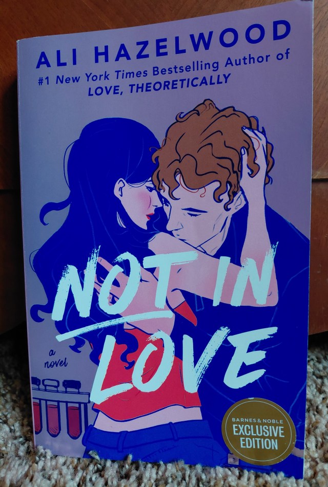 Image is a book cover for Not In Love by Ali Hazelwood. It shows a woman embracing a man. She has bright red lips, black hair, and pale skin. The man has orange curly hair. There are test tubes in the background. 