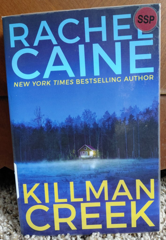 Image is a photo of a book called Killman Creek by Rachel Caine. It's blue and it shows a dark forest with a cabin below it. There's a single light shining on the cabin. It appears that the cabin is on a swamp. 
