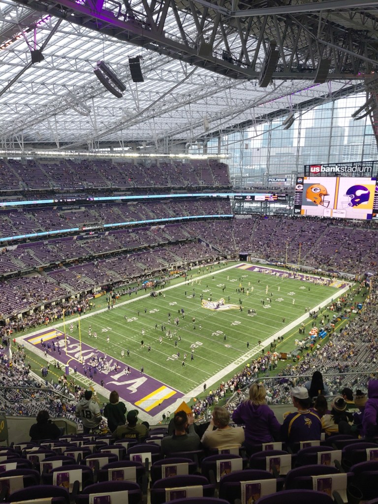 Image is a photo taken from US Bank Stadium. Behind the sign for US Bank Stadium is the Minneapolis skyline. It shows the stadium with both ends saying Vikings in purple and yellow. There are players on the field. 