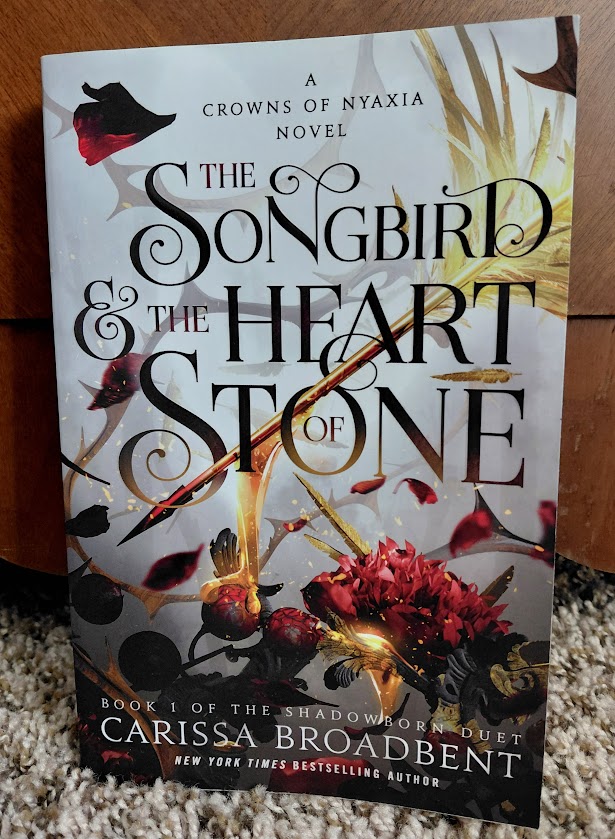 Image is a photo of a book cover for The Songbird & the Heart of Stone by Carissa Broadbent. On the cover, it shows a golden arrow with a yellow feather at the end. It's dripping what appears to be honey or a similar consistency onto a red flower. The background is a bunch of thorny branches in gray mist. 