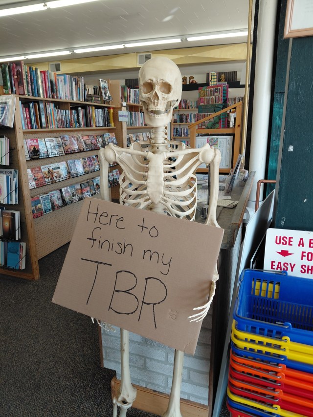 Image is a photo of an anatomy skeleton. The skeleton is holding a cardboard sign and it reads "Here to finish my TBR" 