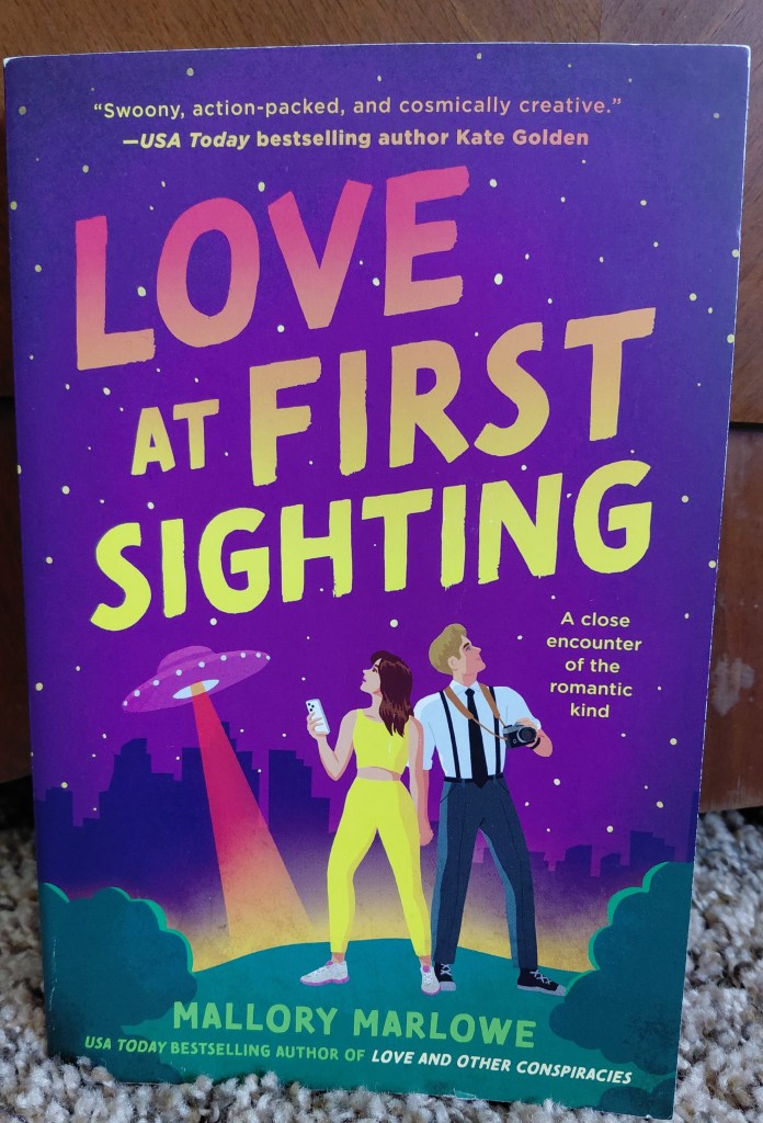Image is a photo of a book called Love at First Sighting by Mallory Marlowe. The sky is a purple pink color and there's a UFO in the background shining a red beam. There's a woman dressed in matching yellow athletic clothing staring at the sky. To her right is a man in a white dress up shirt, black tie and pants holding a camera. He's also looking at the sky. 