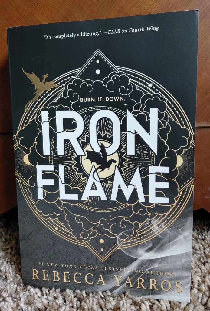 Image is a physical book of Iron Flame by Rebecca Yarros. It is a black and gold book cover. It shows a circle with various lines and suns in the border. There are black clouds outlined in gold. In the center is a gold circle with a black dragon on it. Towards the bottom of the book are white, whispy fog. 