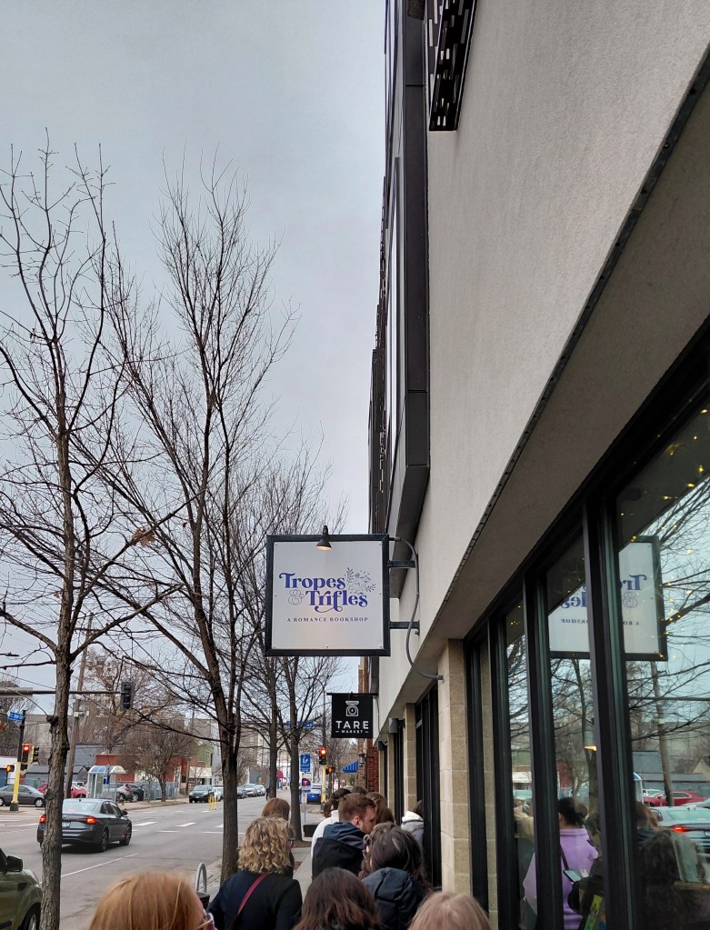 Image is a photo taken outside of a bookstore, Tropes & Trifles. The sign is outlined in black, with a white background and the logo Tropes & Trifles is purple. Outside the store are people waiting to get inside. 