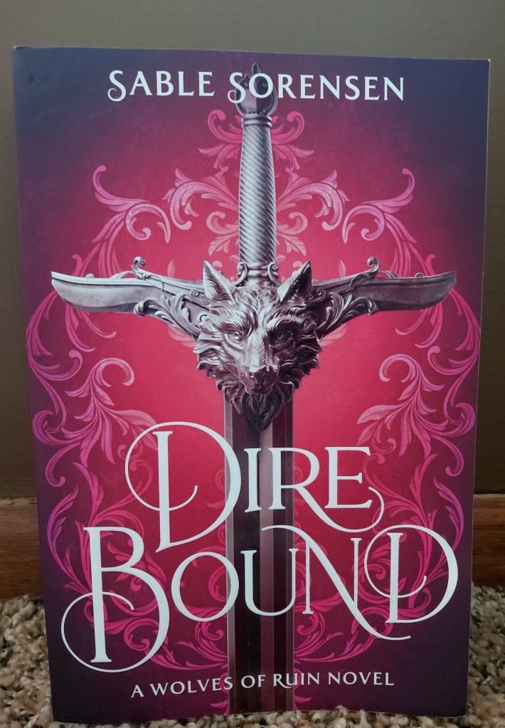 Image is a book cover for the book Dire Bound. The cover is a magenta and purple cover. There are pink what appears to be branches or curls that wrap around in a circle. In the middle is a big gray sword and at the center is an intricate wolf. 