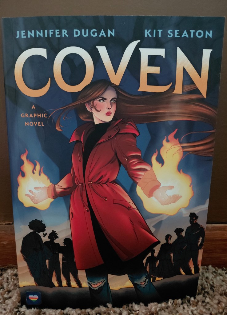 Image is for the graphic novel Coven. It shows a woman with brown hair, wearing a red trenchcoat. In both hands, she has flames. There are black shadowy figures behind her.