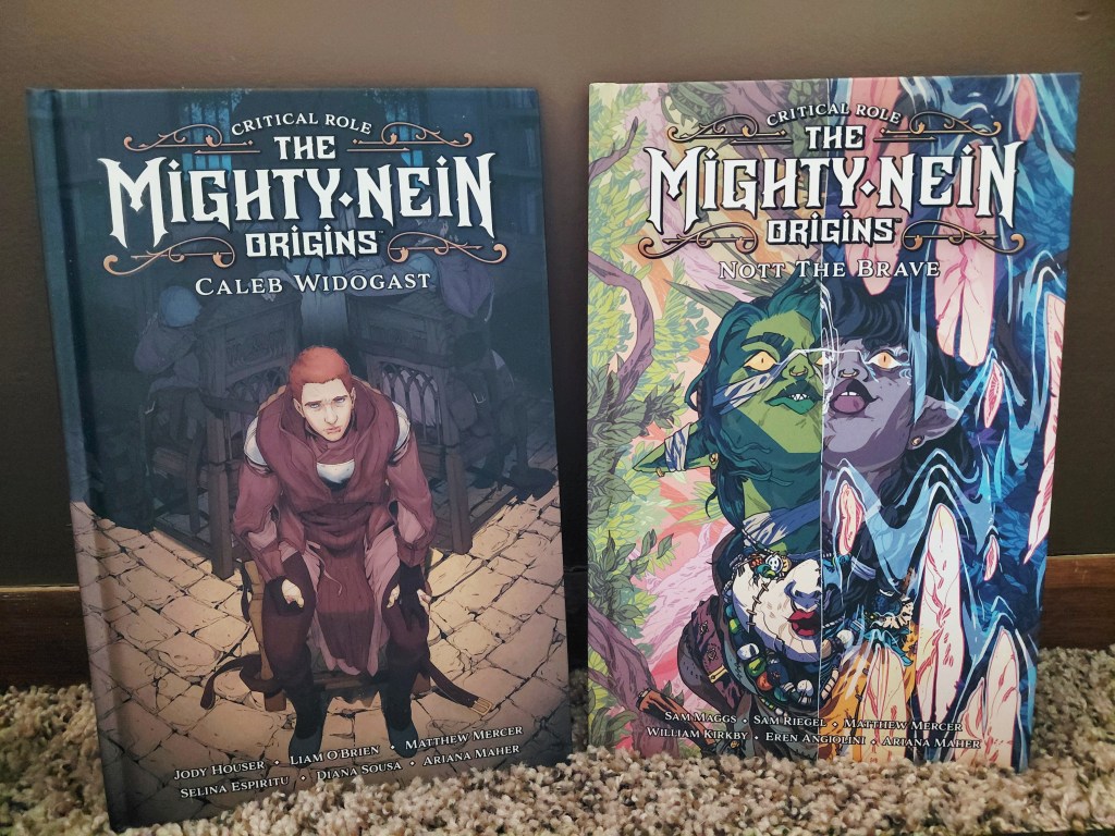 Image is a photo of two graphic novels. The Mighty Nein Origins Cale Widogast and The Mighty Nein Origins Nott The Brave. For the Caleb cover, it shows a young man sitting in a chair, he has a look of anguish or fear on his face. On the Nott The Brave cover, it shows two images split vertically. On the left is a goblin, on the right, it's a purple skinned figure. The cover is a mixture of greens and blues.