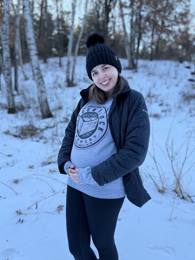 Image is a photo of a woman in black leggings and a gray sweatshirt. It's winter, there's snow on the ground. She's smiling at the camera and her hands are on her lower abdomen. There is a bump there as she's about 20 weeks when this photo was taken. 